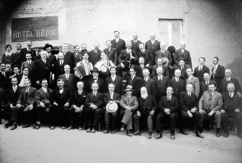 Elus (elegits) et Laguiolais avec accordéoniste (acordeonista) et joueur de cabrette (cabretaires) devant enseigne de l'hôtel Régis, 1931-1932