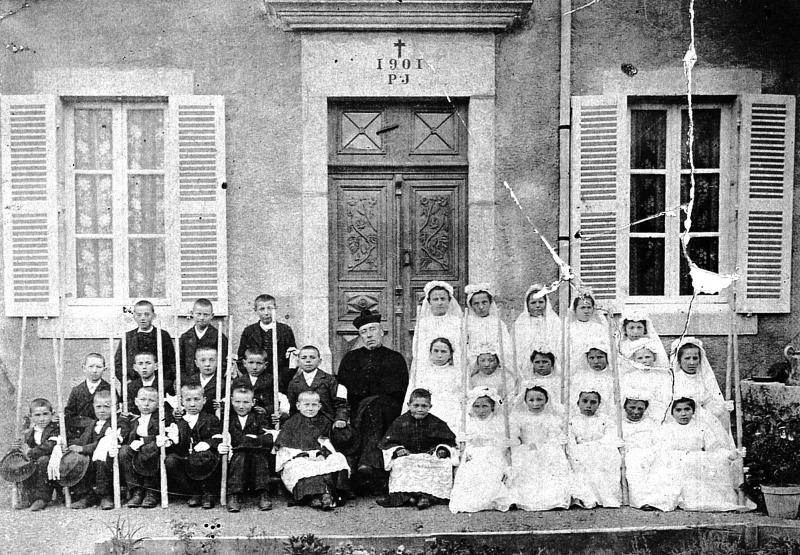 Curé (curat), clercs (clergues), communiants et communiantes en aubes devant porte d'entrée du presbytère (caminada), 1925