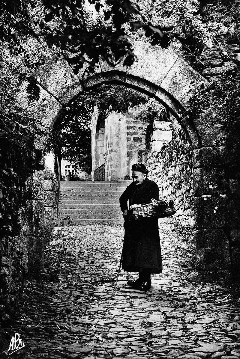 Femme revenant du jardin (òrt) avec panier (panièr) au bras devant porte fortifiée, à Orlhaguet, 1959
