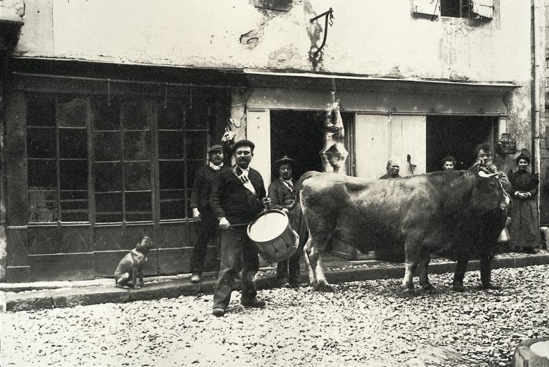 Personnes, joueur de tambour (tamborinaire) et bovidé de race aubrac devant un abattoir (masèl), rue Cornebariols