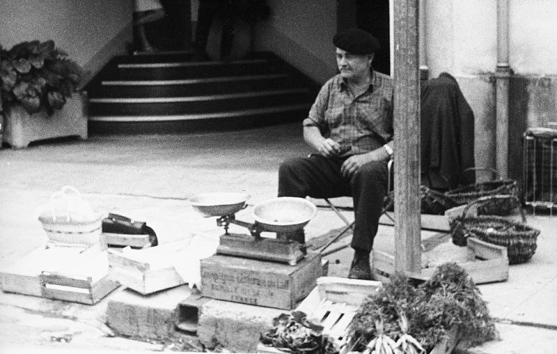 Etal d'une marchande (merchanda) de fleurs et légumes et clientes dans une rue (carrièira) un jour de foire (fièira) ou de marché (mercat), 1969
