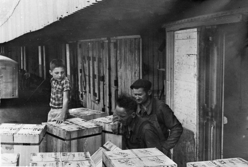 Enfant et deux hommes avec des caisses (gagets) de pains affinés de roquefort devant des containers d'expédition, à la gare