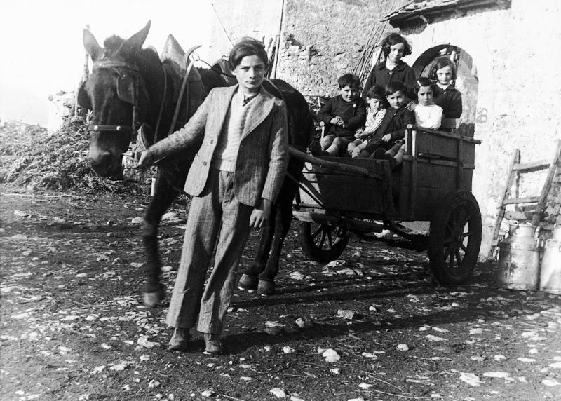 ​Garçon conduisant un équidé attelé à un petit char (carreton) transportant sept enfants devant un portail (portal) de cour de ferme (bòria), aux Fournials, 1935-1940