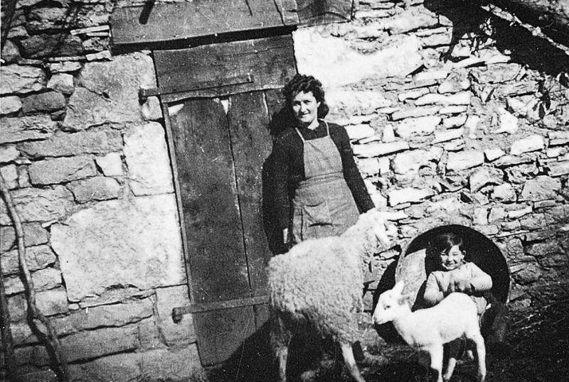 ​Femme avec un garçon dans un baquet, brebis (feda) et son agneau (anhèl) devant une porte d'étable (estable), au Mas de Combettes