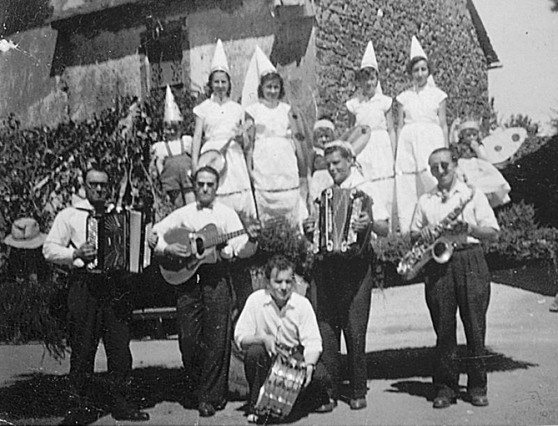 Cinq musiciens (musicaires) d'une fanfare, jeunes filles déguisées en fées (fadas) et enfants en insectes sur une remorque un jour de fête (fèsta, vòta), à La Plancade, 1952