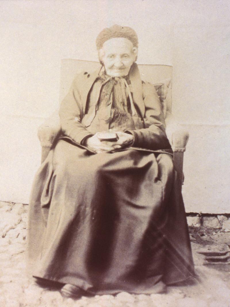 Vieille dame avec coiffe (còfa) et livre dans les mains assise dans un fauteuil, au Bosc (?)