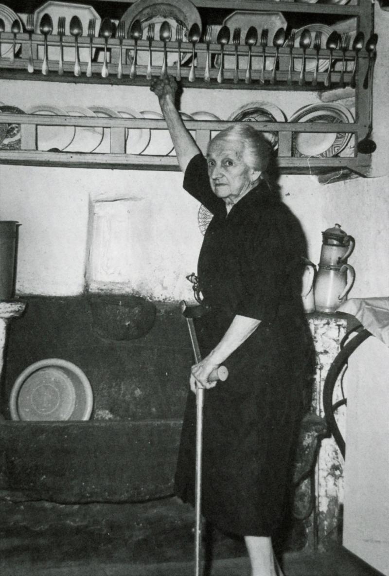 Femme devant un évier (aiguièira) prenant une cuillère (culhièr) dans un vaisselier (drèiçador, vaisselièr), à Serres