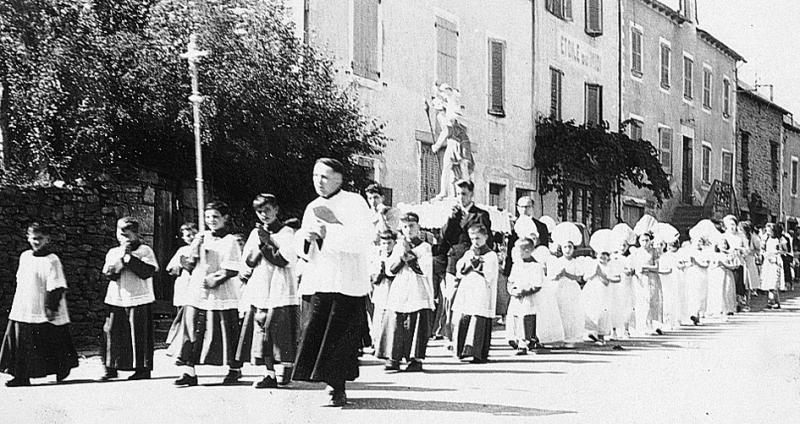 Curé (curat), enfants de chœur (clergues), marguilliers (marguilhièrs) et Enfants de Marie durant la procession de la statue (estatua) de saint Christophe dans une rue (carrièira) pour la Fête Dieu 