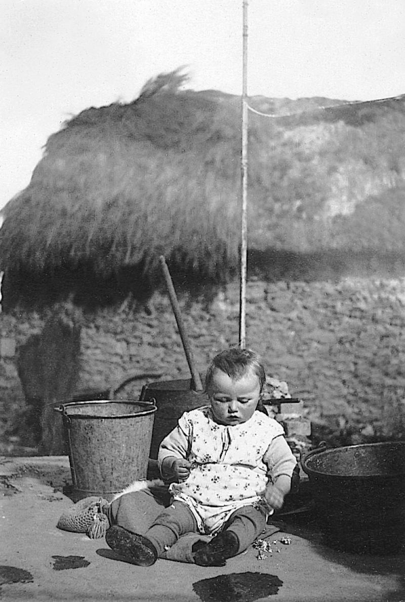 Jeune enfant assis au milieu de seau (farrat), marmite (ola) et petit chaudron (pairolet) devant un bâtiment à la toiture végétale (clujada), à Puech Pouget, 1931