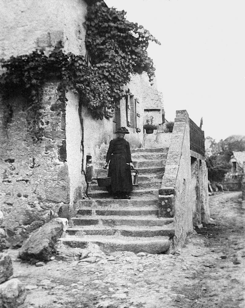 Vieille dame avec un petit chaudron (pairolet) et un seau (farrat) à chaque main dans les escaliers (escalièrs) en pierre d'une maison (ostal), à Douzoulet, 1910