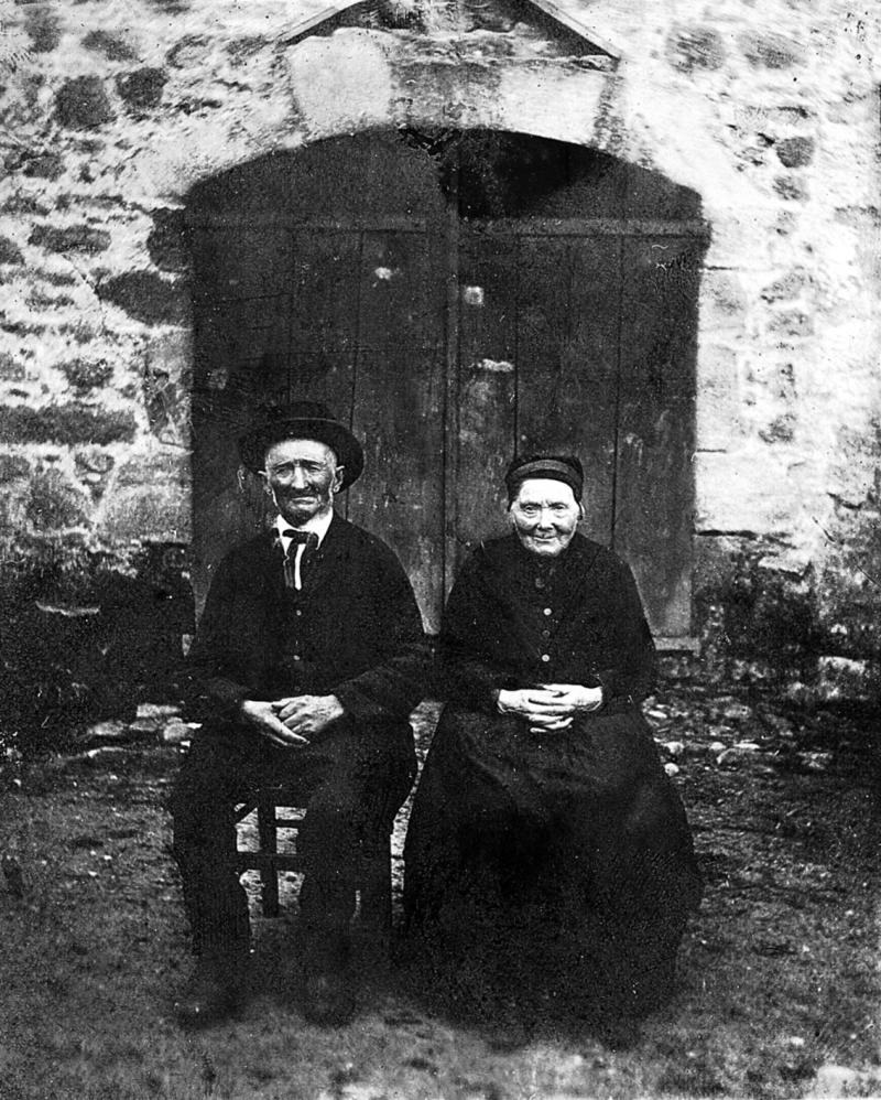 Couple (coble) en habits (vestit) traditionnels assis devant une porte (pòrta) avec triangle de décharge, à Rieupresens