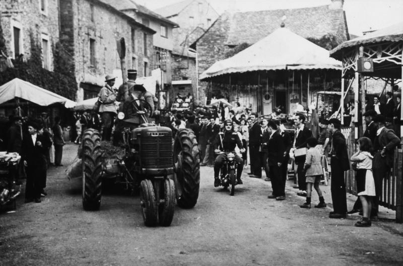 Forains, villageois (vilatjors) regardant passer le char tiré par un tracteur (tractor) d'une cavalcade, manèges (manètges), en Ségala (secteur de Cassagnes Bégonhès ; Salmiech ?)
