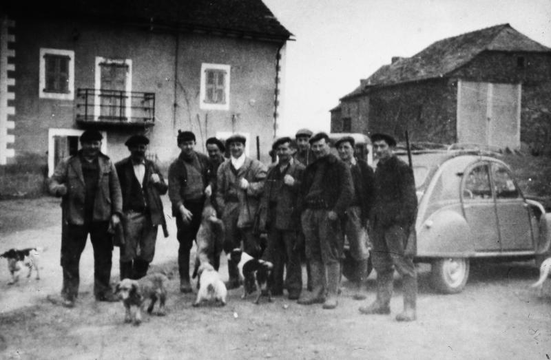Groupe de chasseurs (caçaires), dont un présentant un renard (rainald) à un chien (can), et voitures (veituras) dans une cour de ferme (bòria), en Ségala (secteur de Cassagnes Bégonhès : Cassagnes Bég