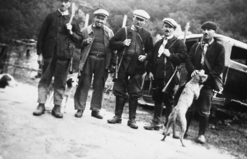 Cinq chasseurs (caçaires) et trois chiens (cans) devant une voiture (veitura) dans une cour de ferme (bòria), en Ségala (secteur de Cassagnes Bégonhès : Cassagnes Bég